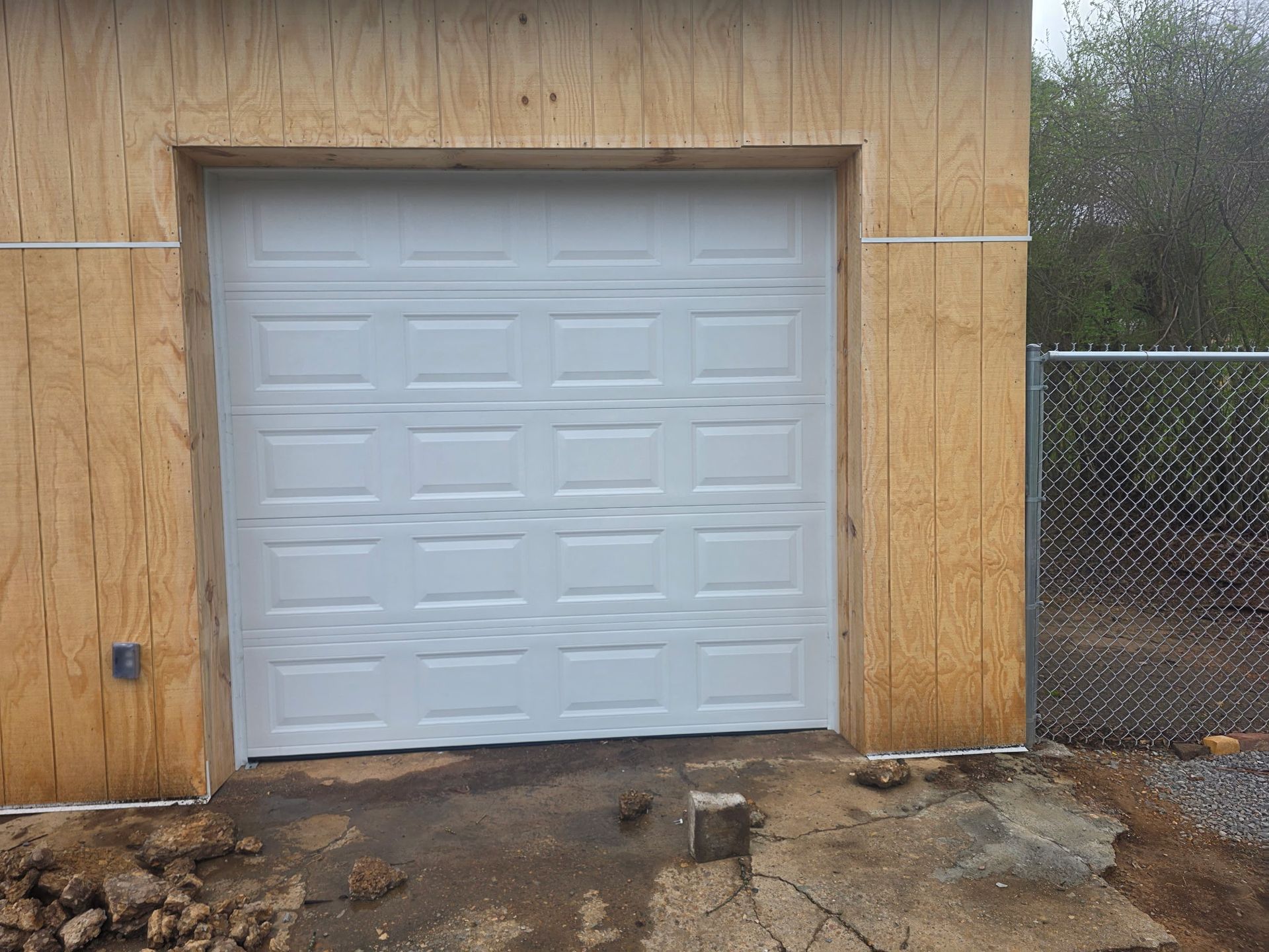 White garage door in a plywood-walled building with muddy ground in front and a chain-link fence on the right.