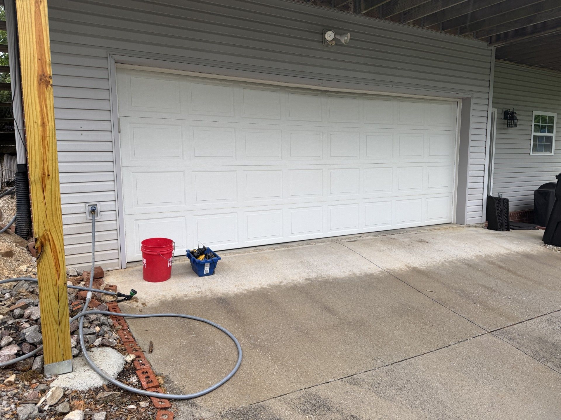 Closed white garage door with red bucket and blue tool on concrete driveway.