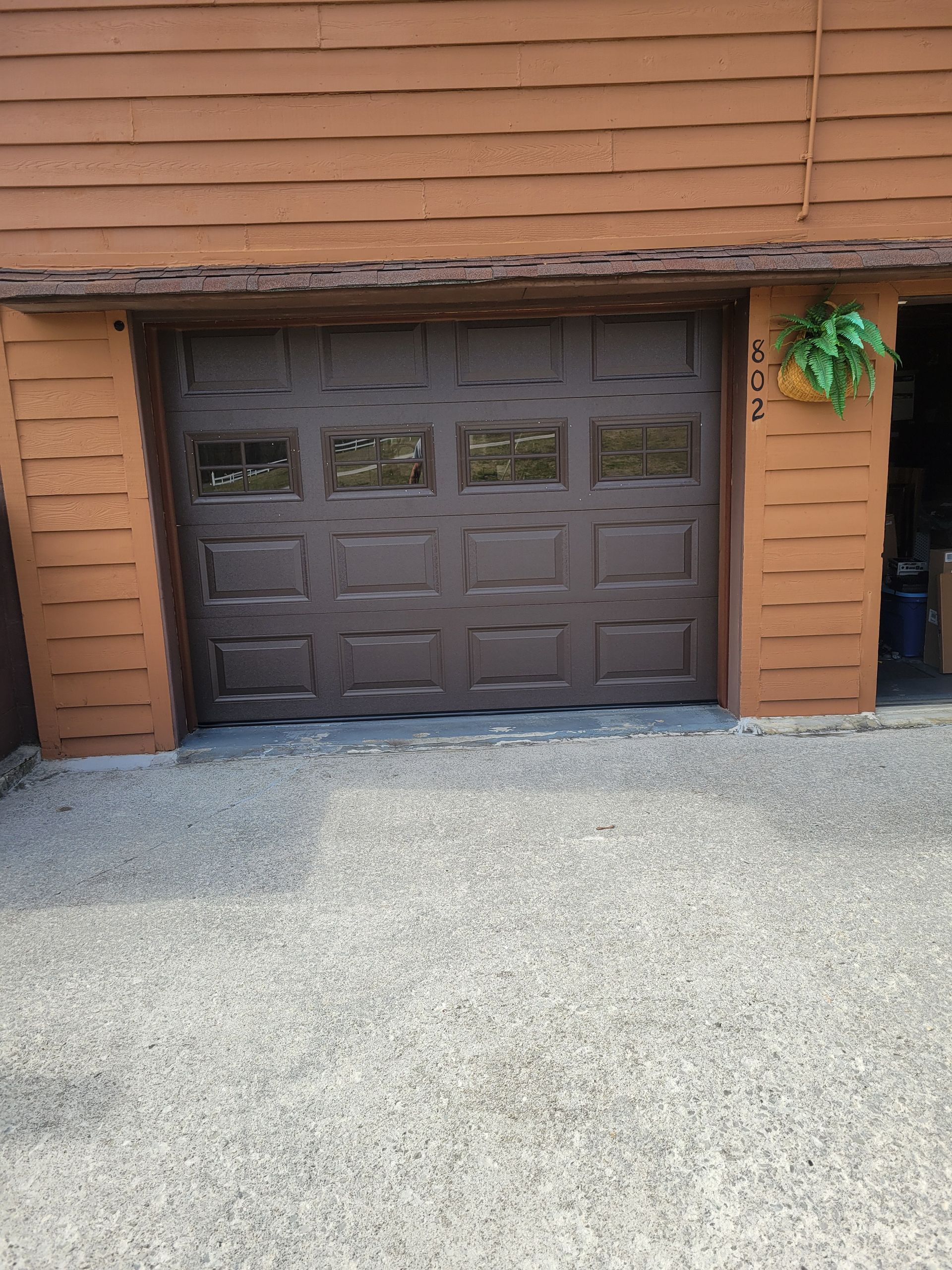 Brown garage door set in a wooden exterior wall with gravel in front and a small hanging plant on the right.
