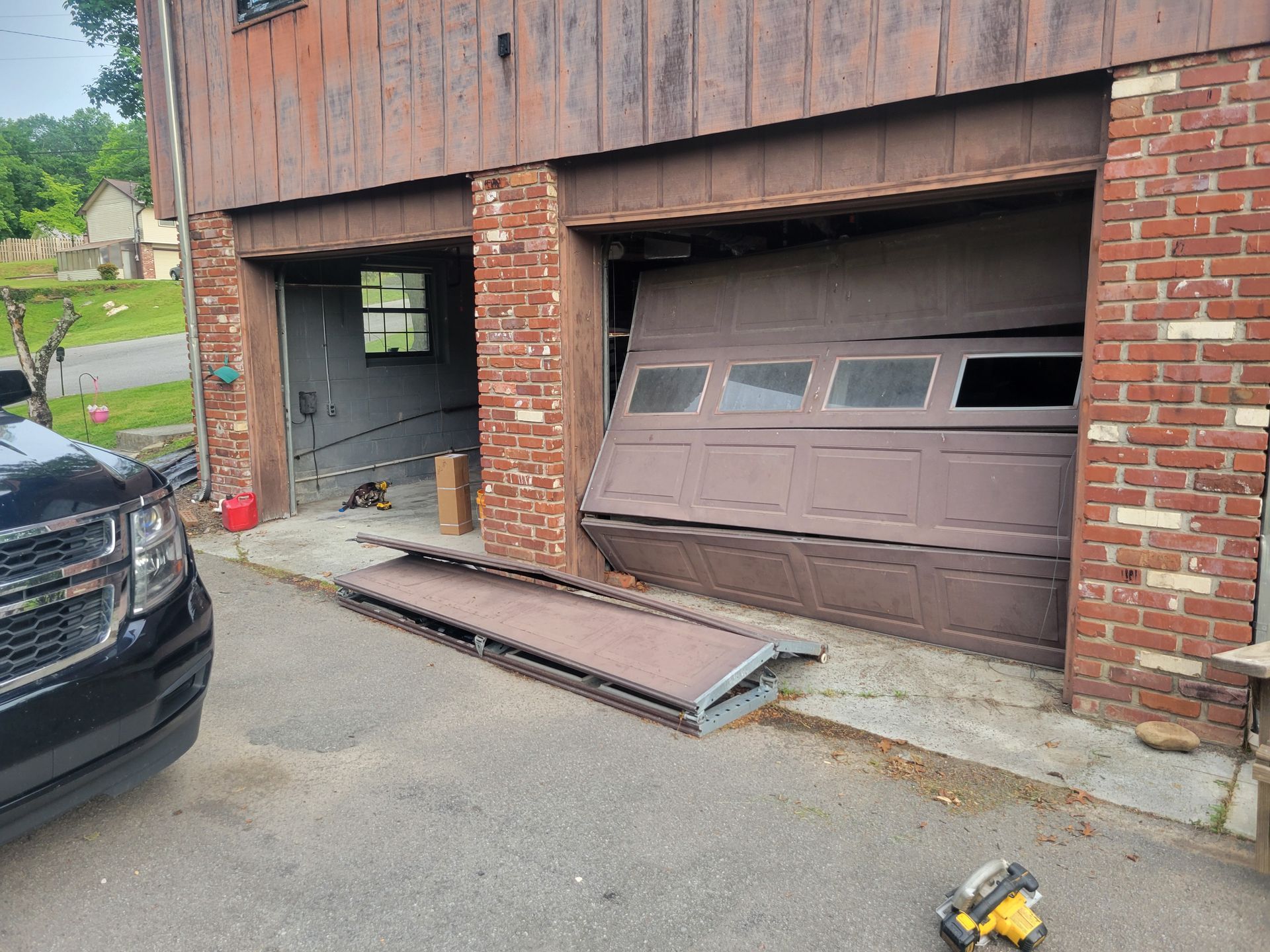 Damaged garage door with a detached panel leaning on the driveway beside a brick building