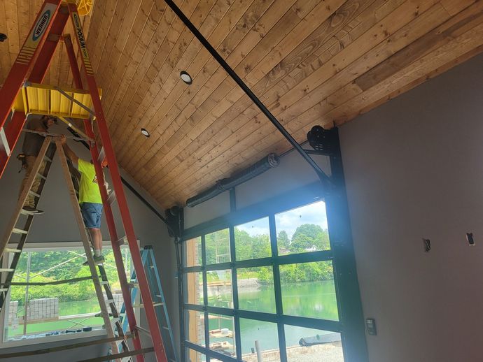 Worker on a red ladder installing a wooden ceiling above large windows in a bright room.