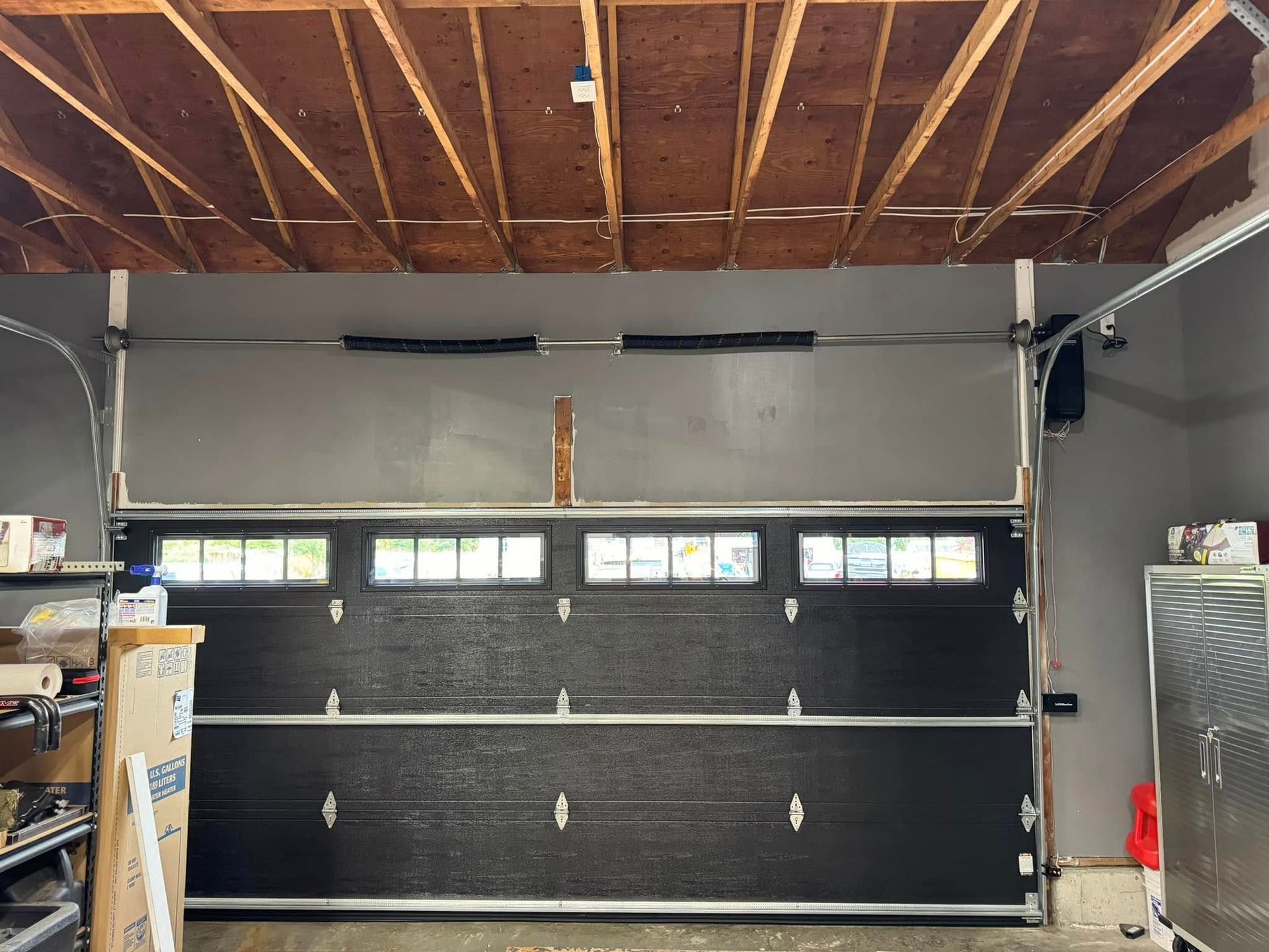Inside a garage, a closed black sectional door sits below small windows and an exposed wooden ceiling.