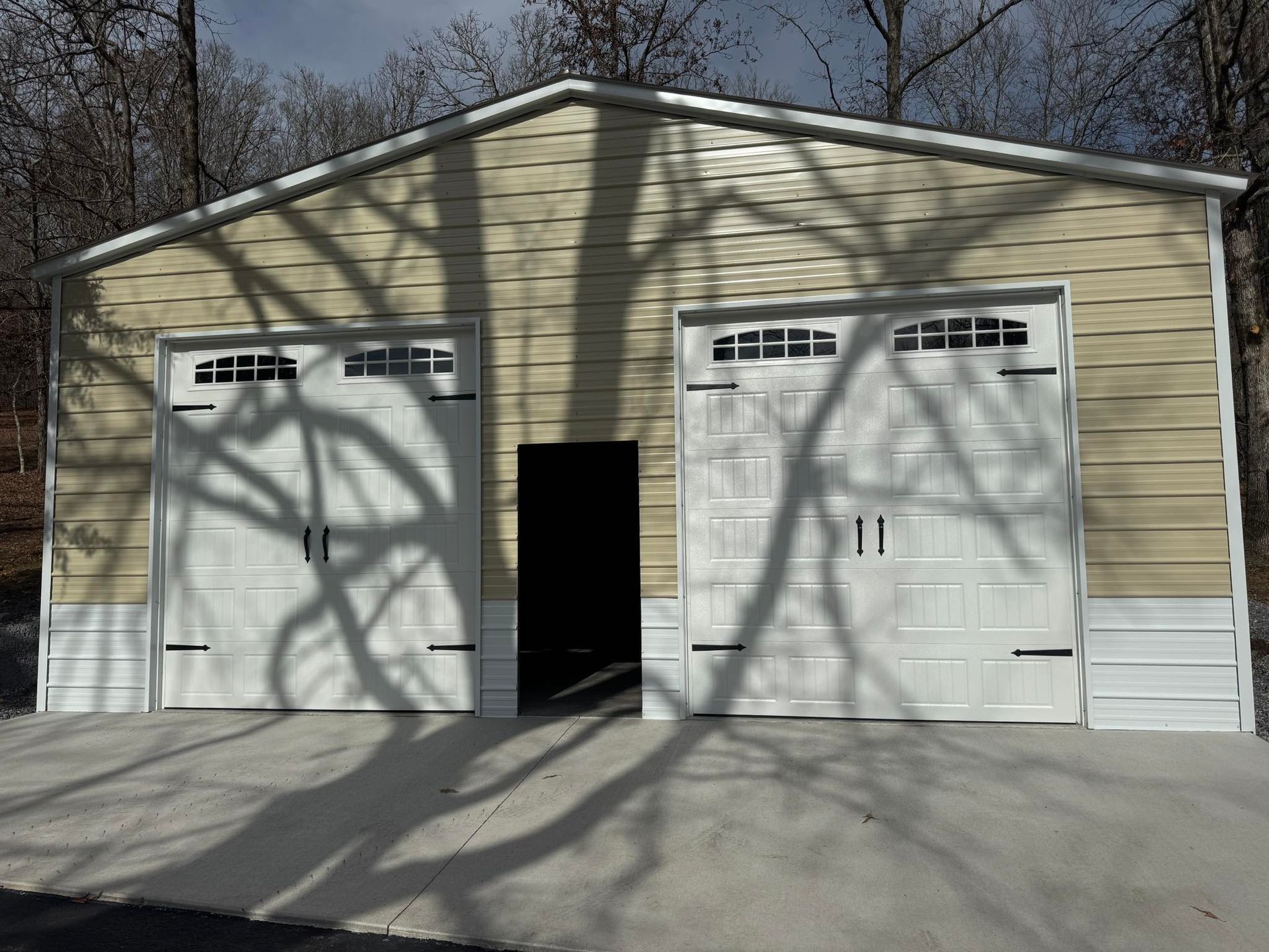 Detached beige garage with two white doors and an open center entry, trees casting shadows across the front.