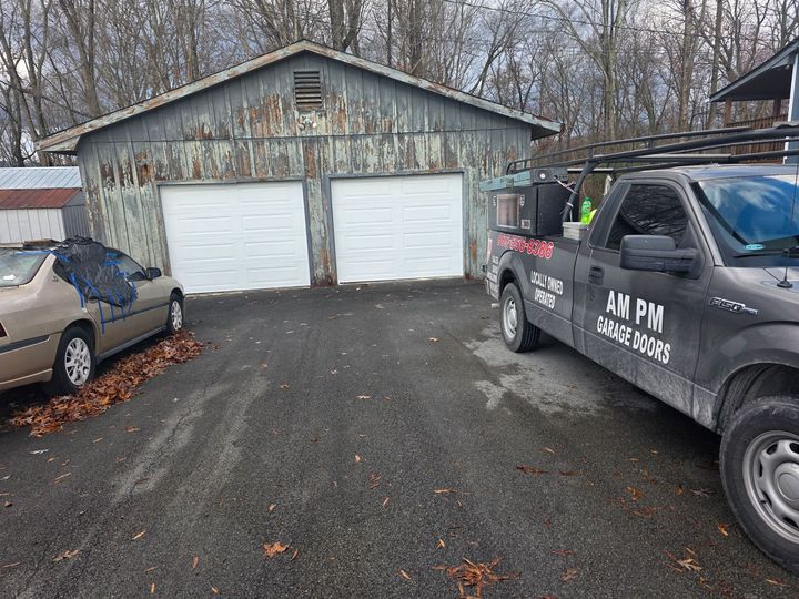Two white garage doors on a weathered shed, with a pickup truck and car parked on a gravel driveway.