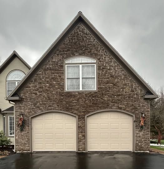 Two-car beige garage with stone facade and white-trimmed gable window under a cloudy sky