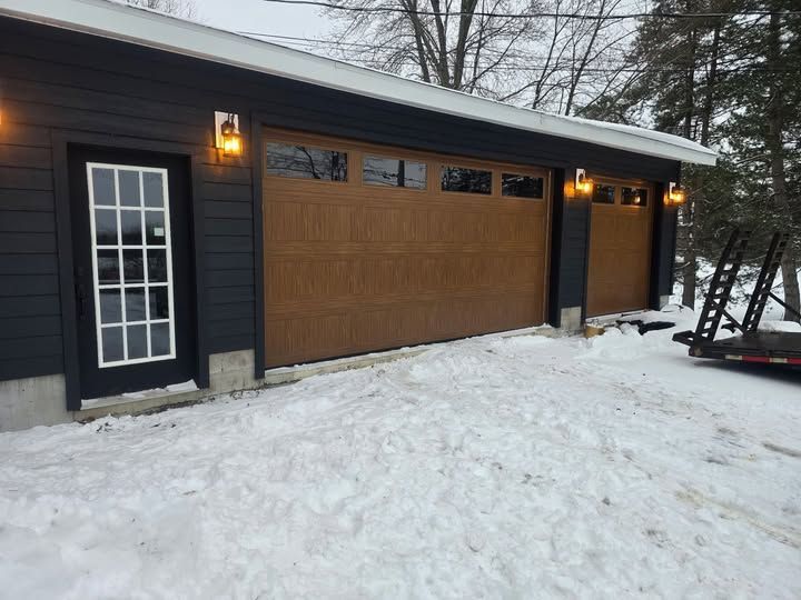 Snow-covered garage with dark siding, wood garage doors, and lit exterior lanterns in winter.