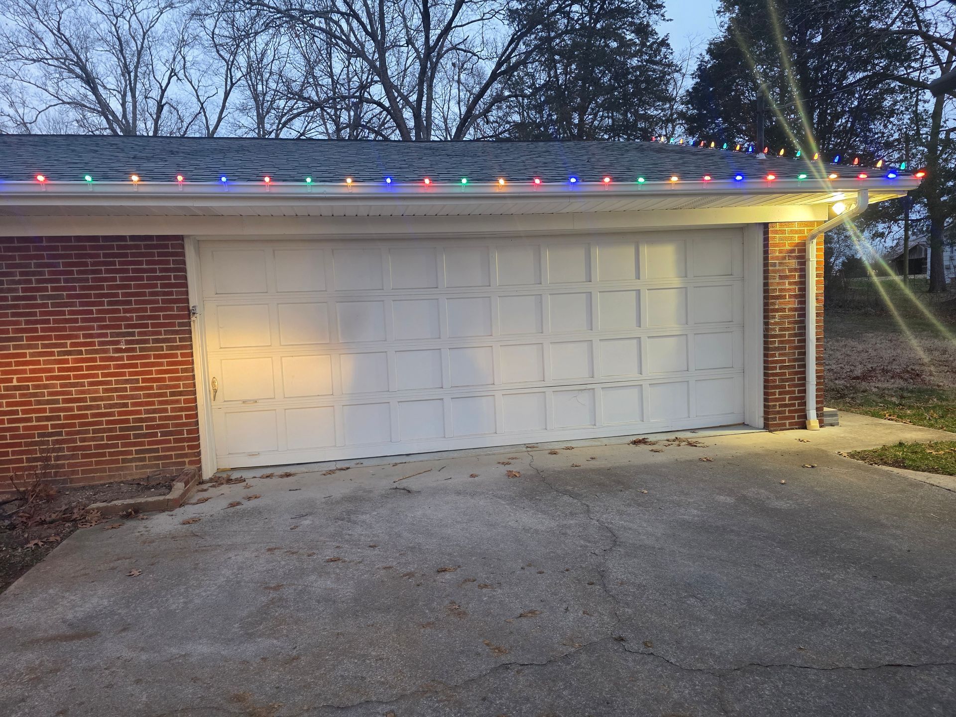 Suburban garage with white door, brick wall, and colorful string lights along the roofline at dusk