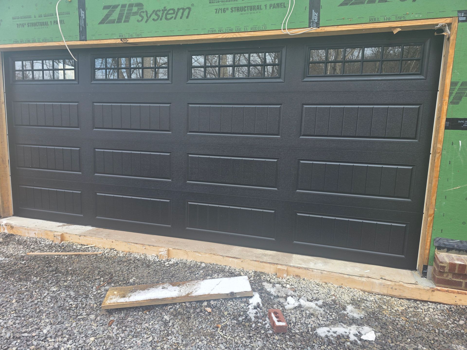 Black garage door with frosted windows in a construction frame, gravel foreground with debris.