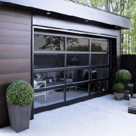 Modern black garage with large glass door, flanked by potted topiary plants on a concrete patio