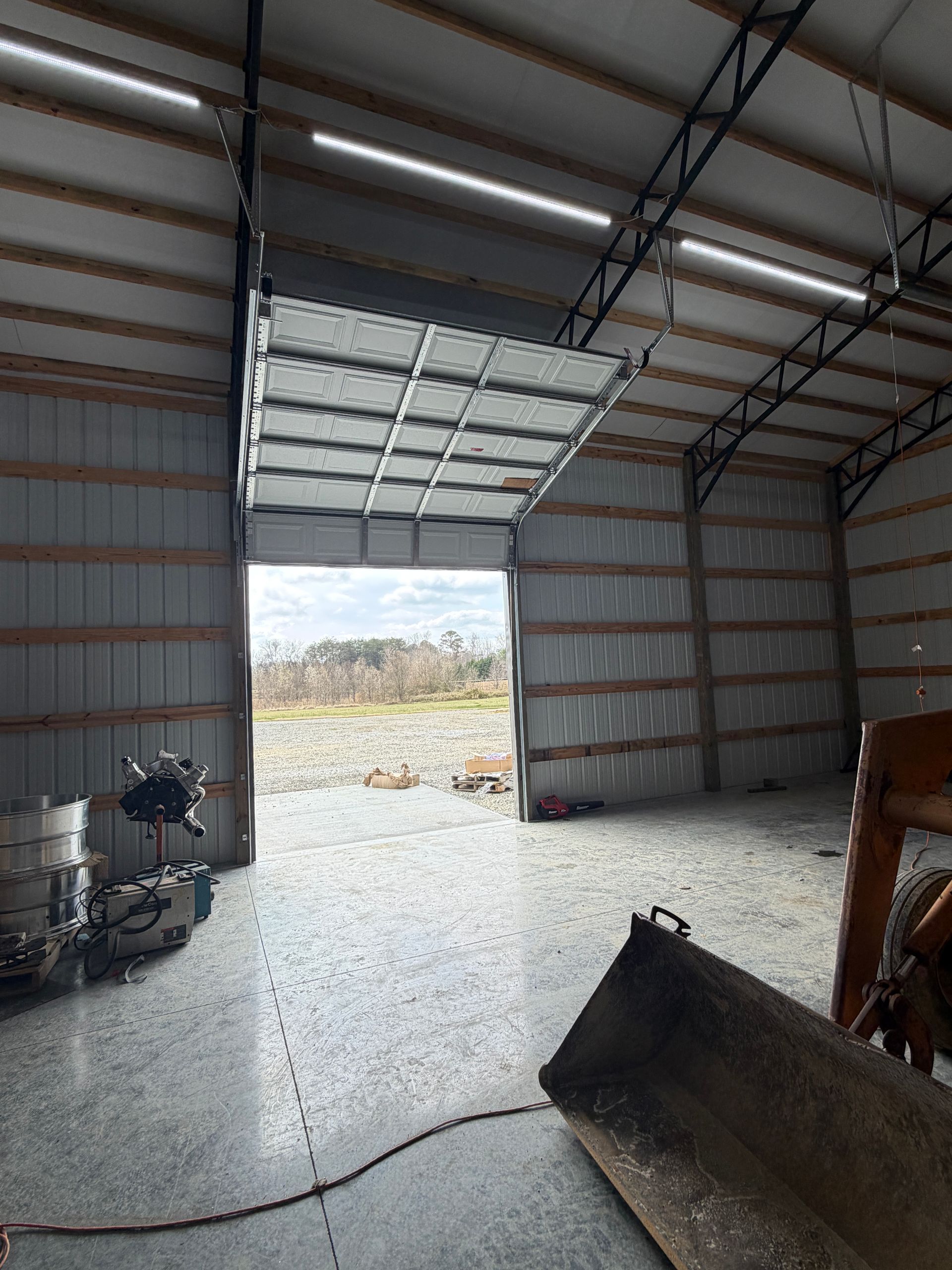 Inside a metal garage, a partially open roll-up door reveals a gravel lot outside.