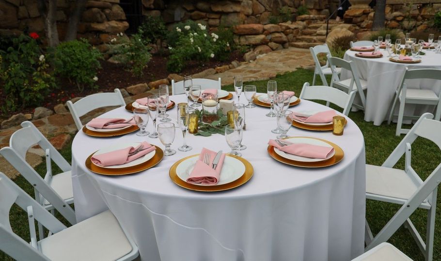 Outdoor round banquet table with white tablecloth, pink napkins, and place settings, surrounded by white chairs.