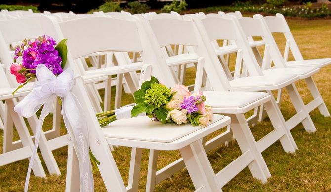 White chairs with purple and pastel flower bouquets arranged outdoors on grass for a wedding ceremony