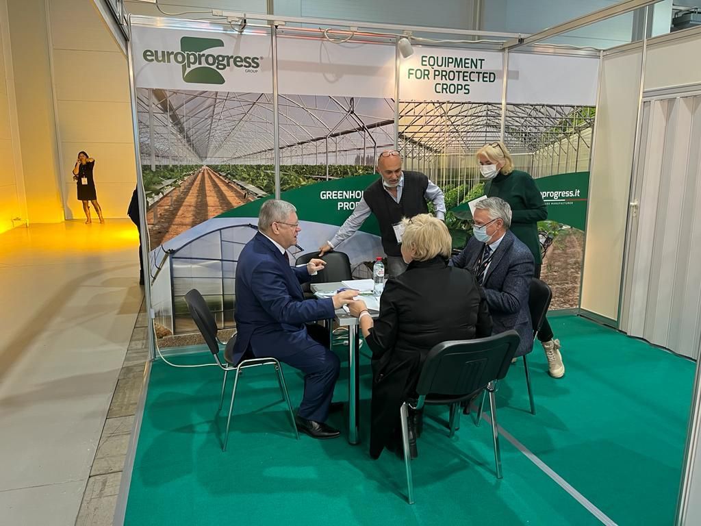 Cinque persone sedute allo stand di una fiera commerciale discutono di un'esposizione di campi agricoli e di una serra.