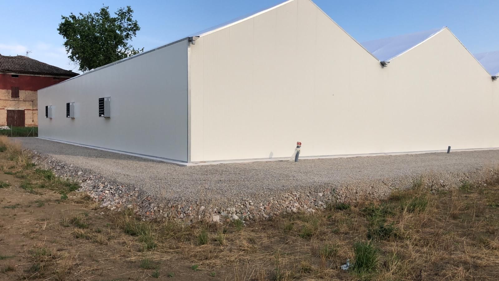 Edificio moderno bianco con tetto spiovente accanto a un cortile di ghiaia e a un prato rado sotto un cielo limpido.