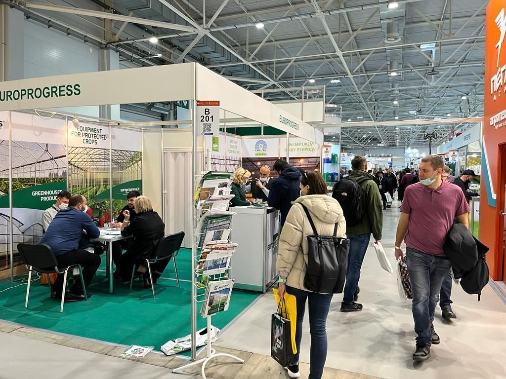Corridoio gremito di una fiera commerciale, con espositori, visitatori seduti e stand sotto un grande padiglione coperto.
