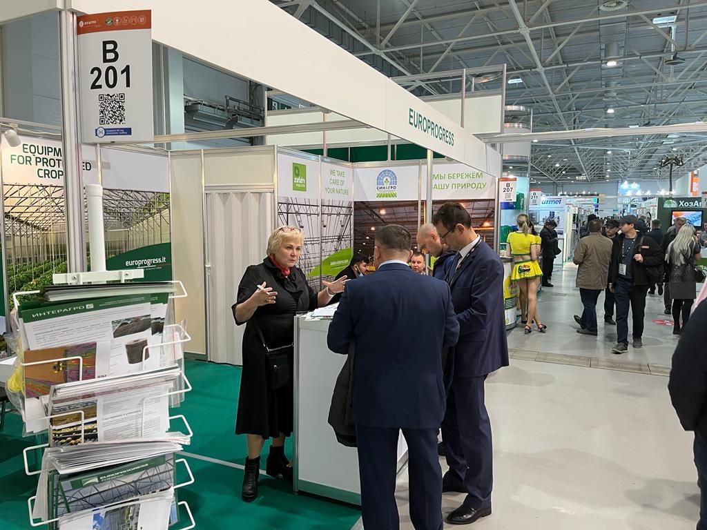 Persone che conversano presso uno stand fieristico con brochure e pannelli espositivi in ​​un ampio padiglione.
