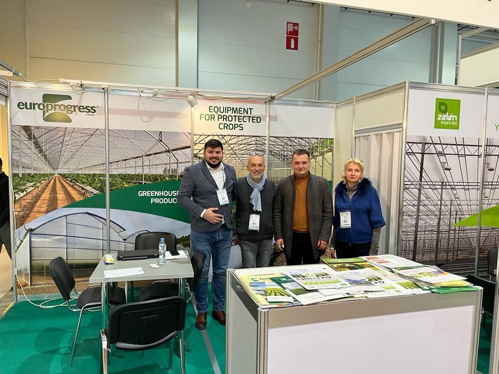 Stand con quattro persone in una fiera, con striscioni verdi e bianchi e un tavolo bianco.