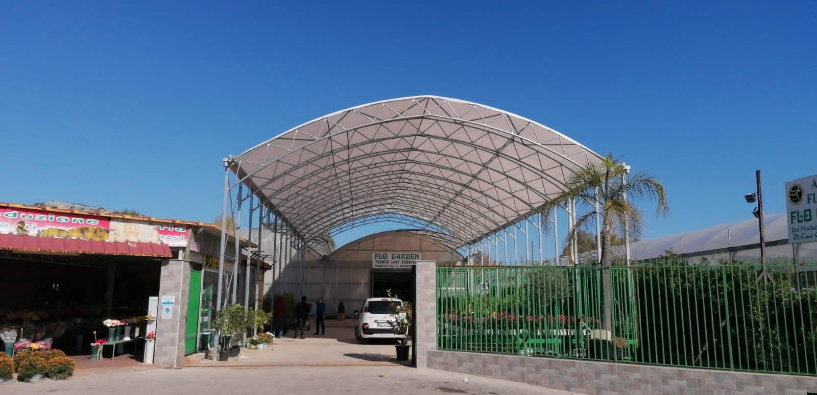 Ingresso con tettoia ad arco sopra un vialetto, recinzione verde e un'auto sotto un cielo azzurro limpido.