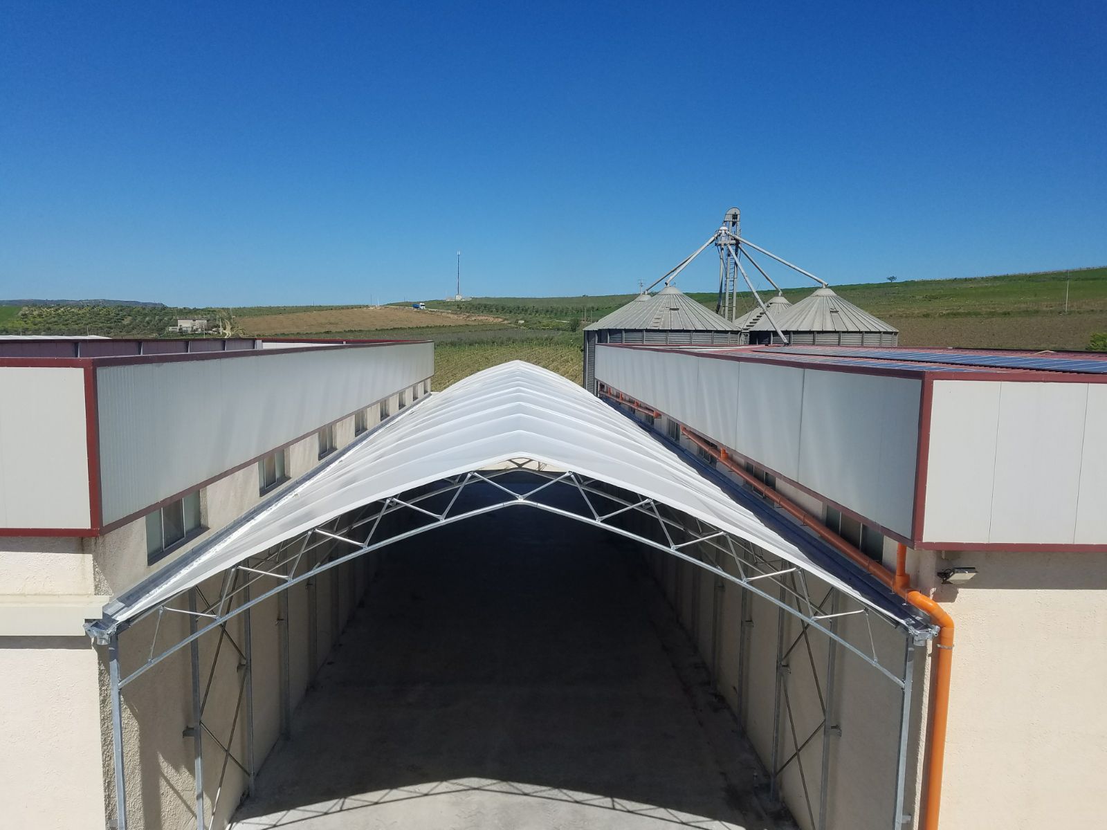 Struttura agricola coperta con tettoia in telone bianco accanto a un deposito di cereali sotto un cielo azzurro.
