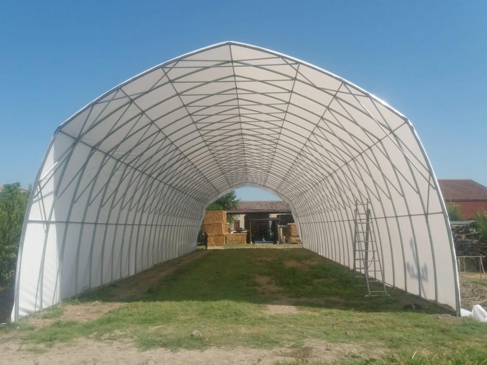 Serra bianca a forma di tunnel, con struttura in legno, aperta su entrambe le estremità, sotto un cielo azzurro, sopra un prato.