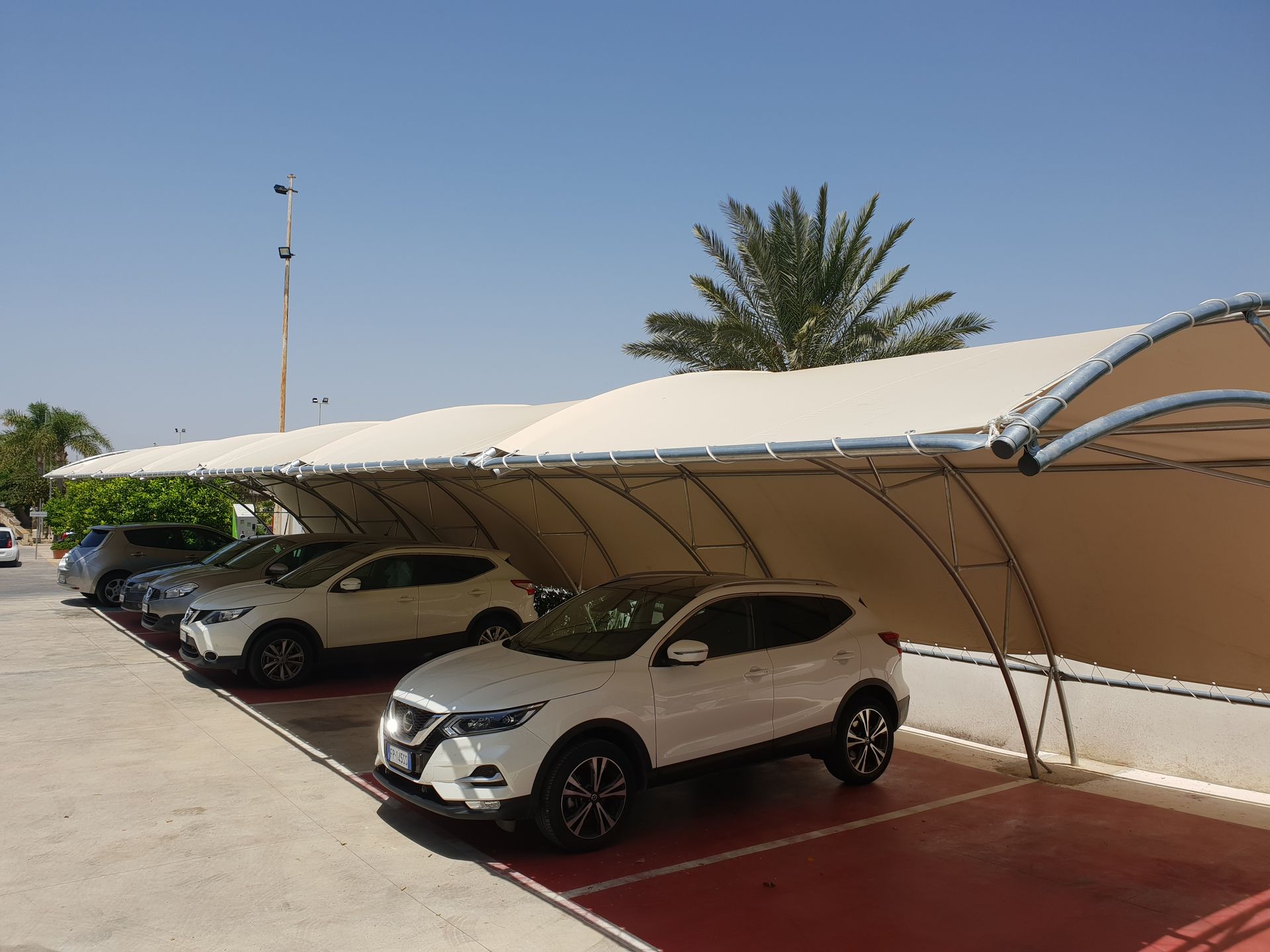 Auto parcheggiate sotto una lunga tettoia bianca in un parcheggio all'aperto in una giornata di sole.