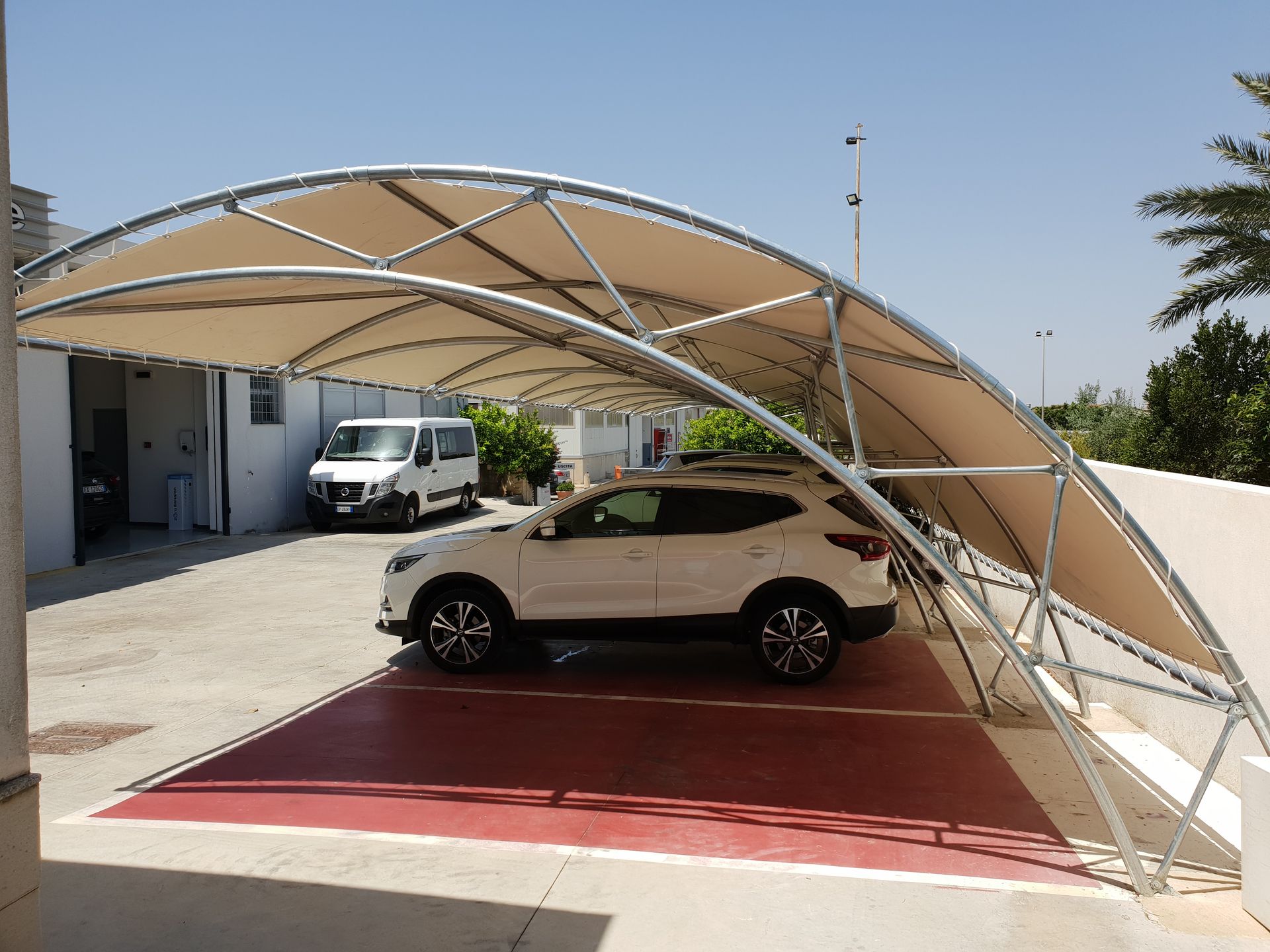 Auto parcheggiata sotto una tettoia beige in un vialetto, con un furgone bianco e un edificio sullo sfondo.