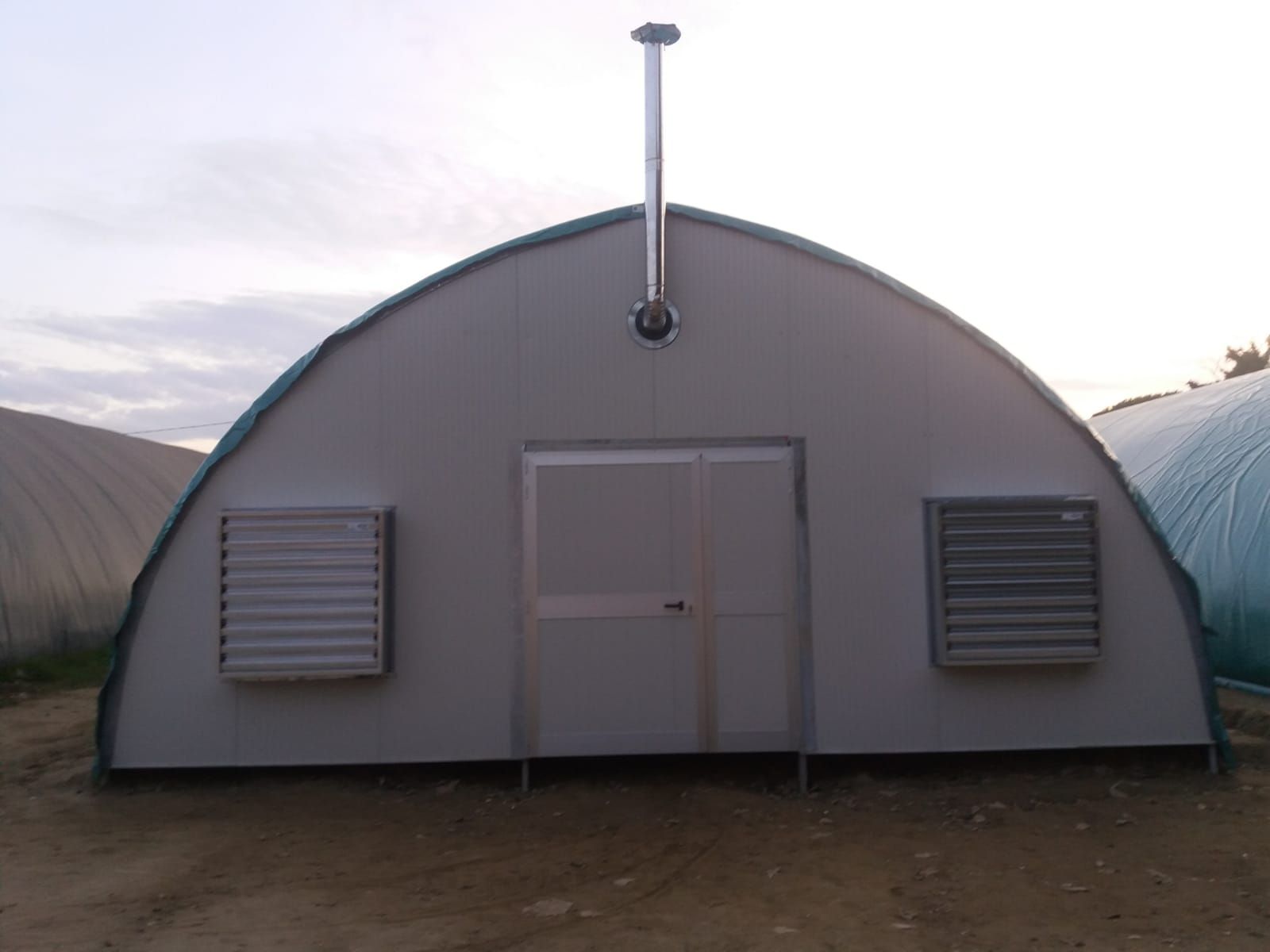 Edificio grigio ad arco simile a una tenda con porta centrale, prese d'aria laterali e camino su un lotto di terra.