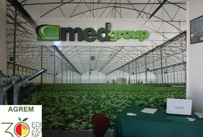 Stand di Medgroup con un grande logo sopra una serra con esposizione di colture verdi e un tavolo antistante.