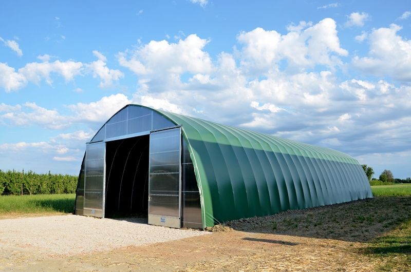 Capanno da giardino verde ad arco con porta aperta in un campo rurale sotto un cielo azzurro nuvoloso.
