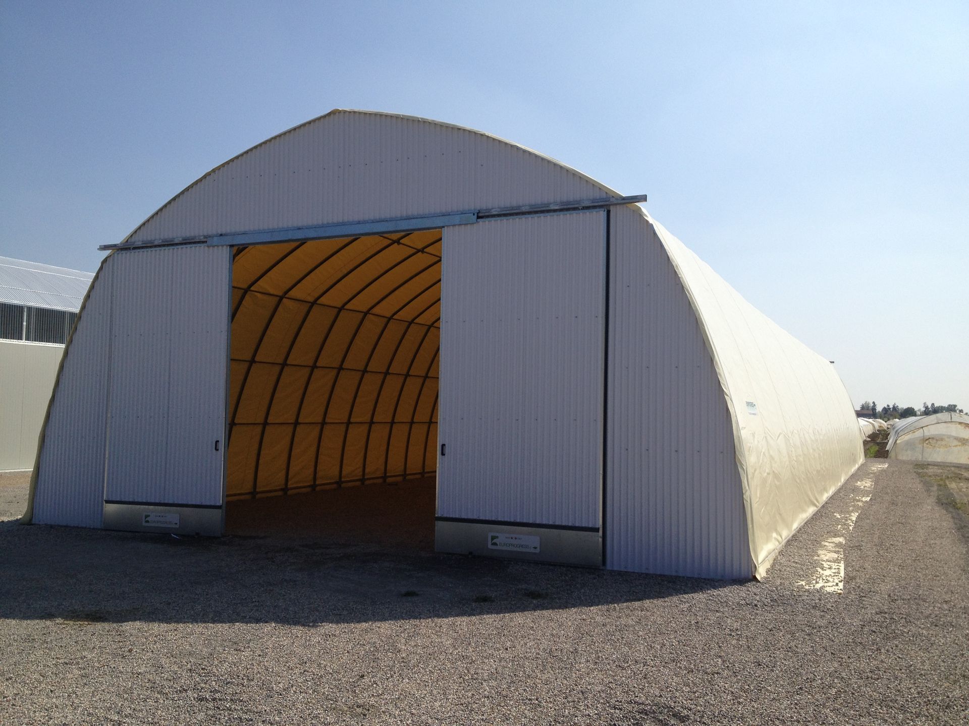 Grande tenda bianca ad arco con ingresso aperto su un piazzale ghiaioso sotto un cielo limpido