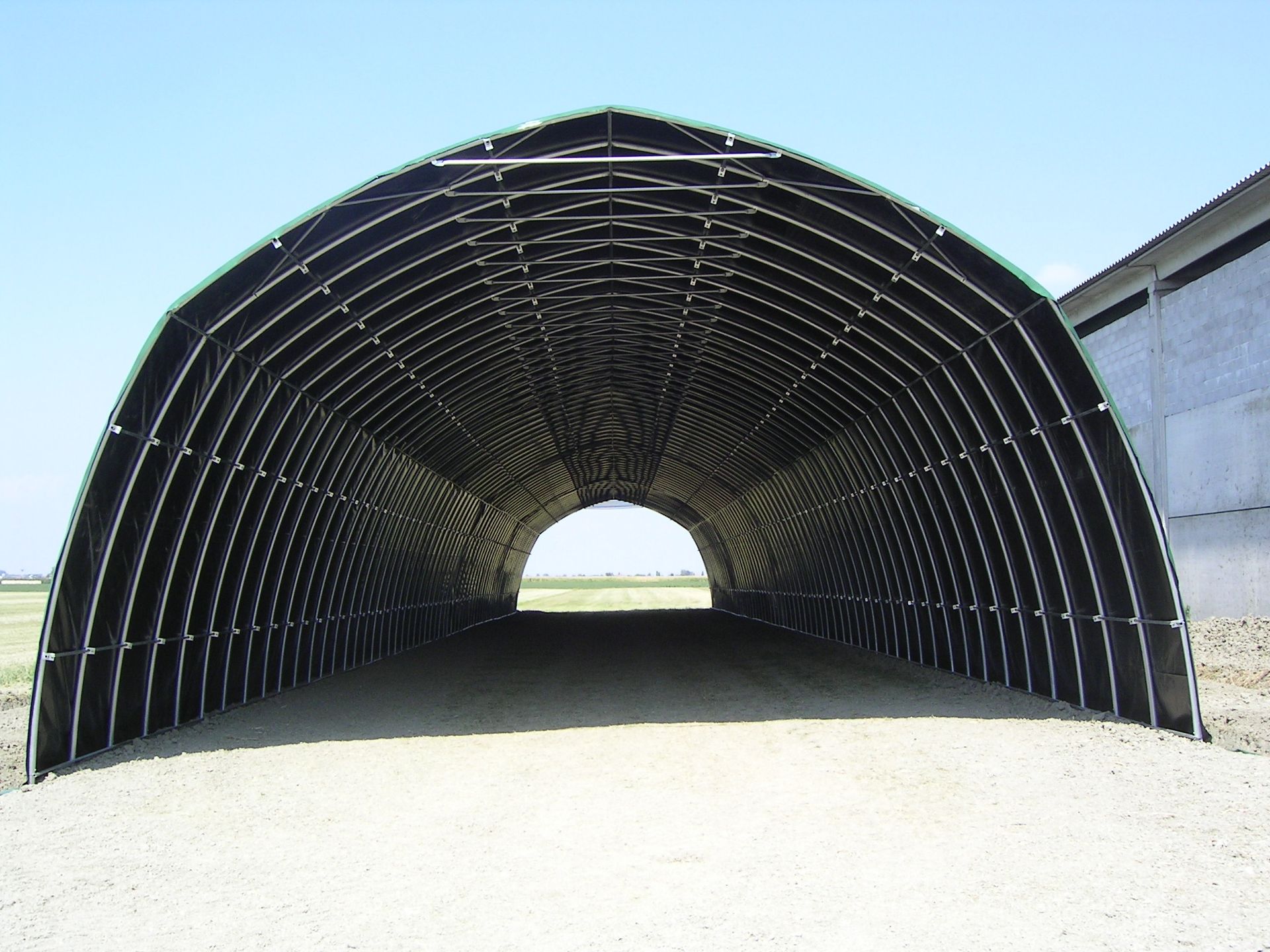 Un grande tunnel metallico ad arco di colore nero, visto dall'ingresso verso l'apertura in una giornata di sole.