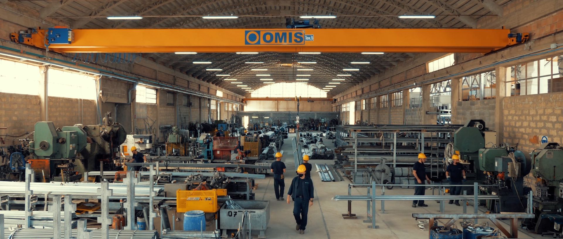 Ampio capannone industriale con operai, macchinari e una gru a ponte arancione all'interno di un grande stabilimento.