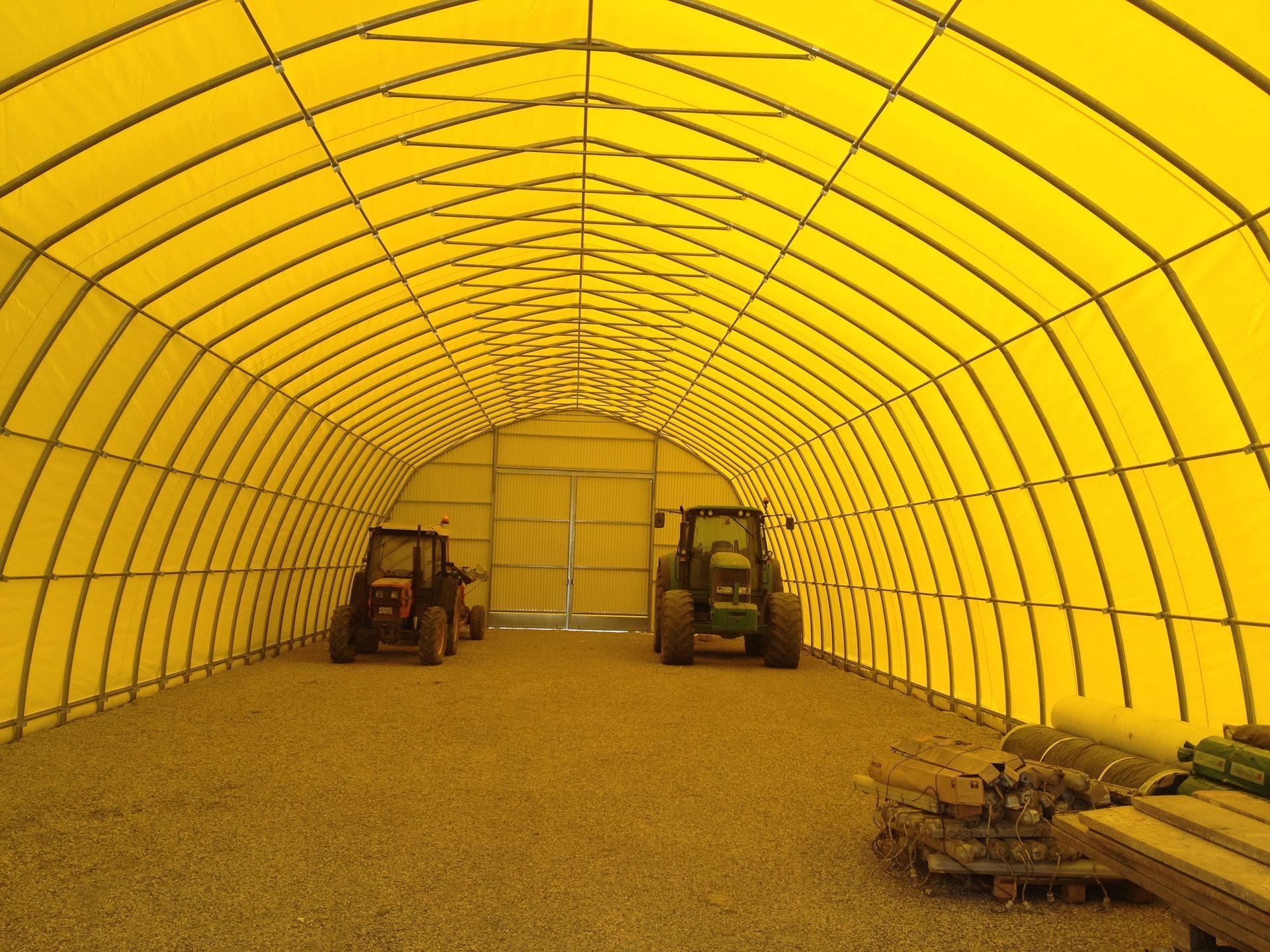 Tenda agricola illuminata di giallo con due carrelli elevatori e materiali impilati all'interno.