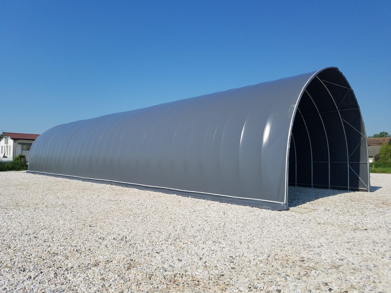 Lungo capannone metallico grigio in stile Quonset su un piazzale ghiaioso sotto un cielo azzurro limpido.
