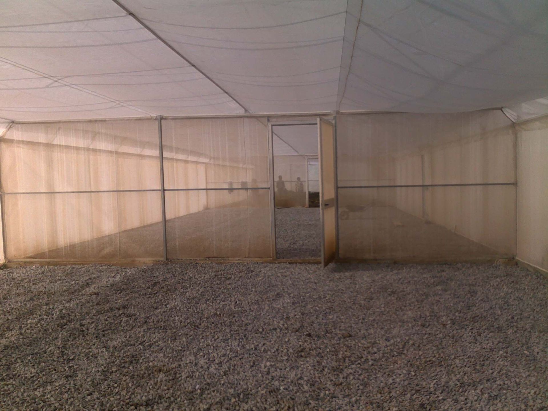 Tenda vuota con pavimento in ghiaia, pareti in rete beige e un'apertura centrale che si affaccia su una vista esterna in lontananza.