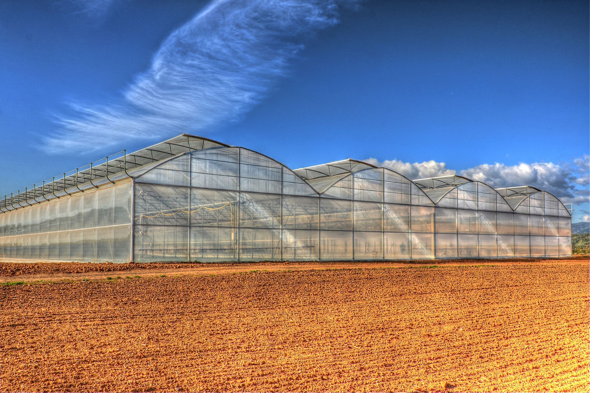 Struttura di serra parzialmente assemblata su un campo di terra battuta sotto un cielo azzurro.