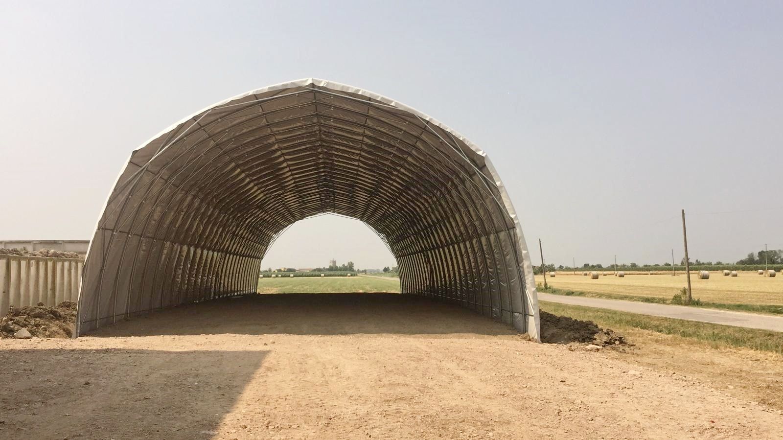 Ampia struttura intrecciata a forma di tunnel che sovrasta una strada sterrata in un paesaggio rurale pianeggiante.