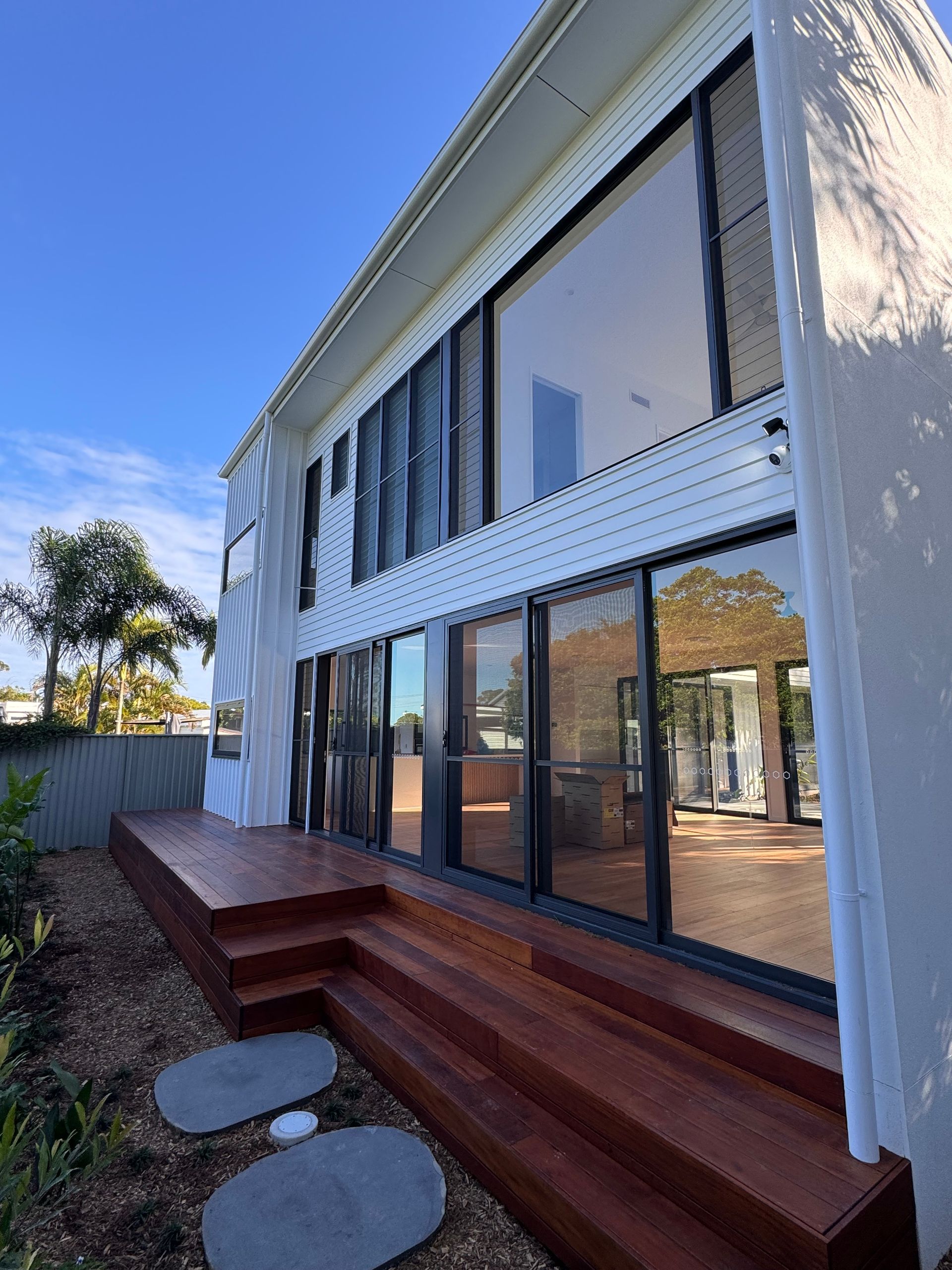 A two-story white building with large floor-to-ceiling windows, featuring a multi-level wooden deck and stone pavers.