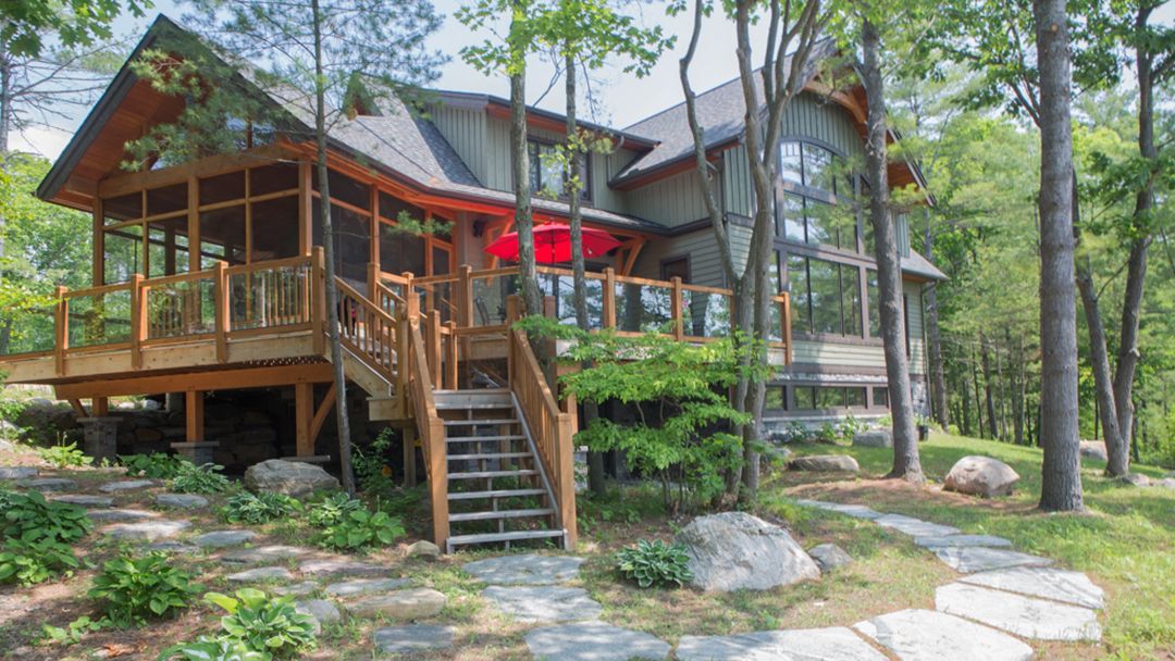 Wooden deck around the Muskoika room of a traditional cottage. Landscaping with walkways and exposed rocks complete the picture.
