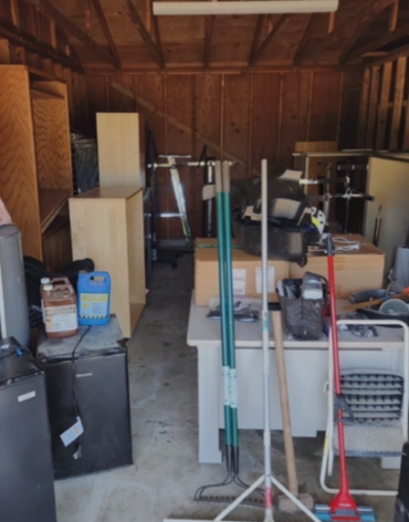Cluttered wooden shed with stacked boxes, tools, and household items on the floor and shelves.