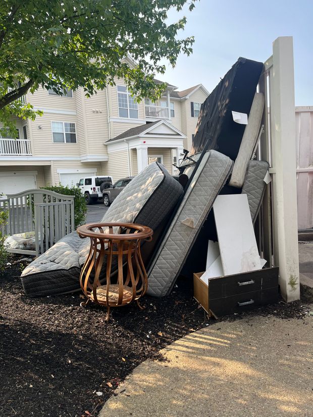 Pile of discarded furniture and debris stacked outside a house along a walkway, with trees overhead.