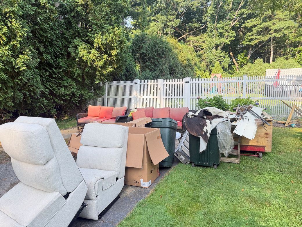 Piled curbside furniture and trash by a white fence in a grassy yard