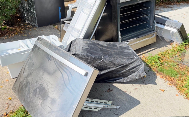 Discarded HVAC unit parts and panels piled beside a sidewalk, with a black trash bag on the ground.