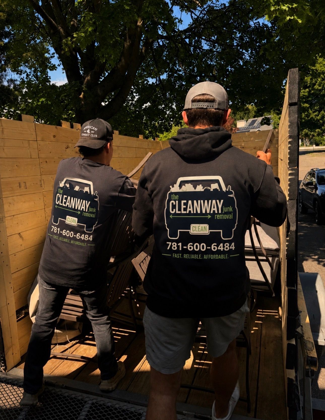 Two workers in black shirts unload equipment outdoors beside a truck and brick wall.