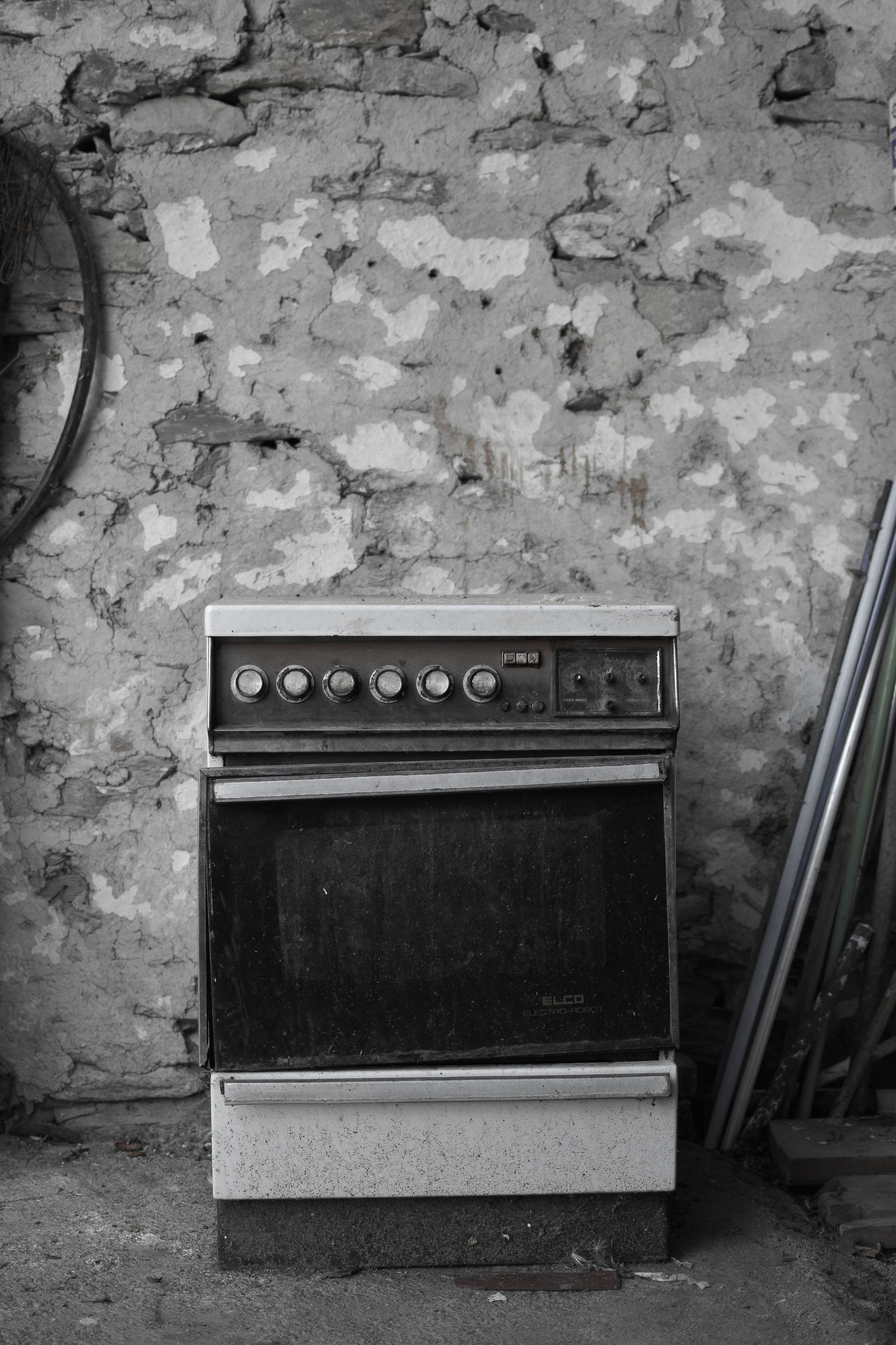 Old stove against a rough stone wall in a rustic room, with tools leaning nearby.