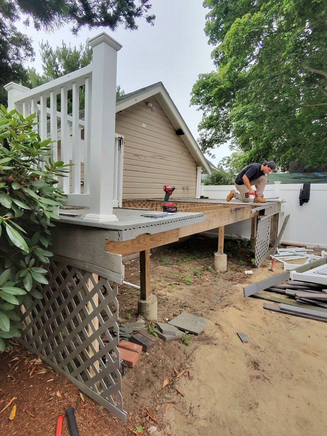 Worker building a raised wooden deck beside a house, with lattice and trees around the yard