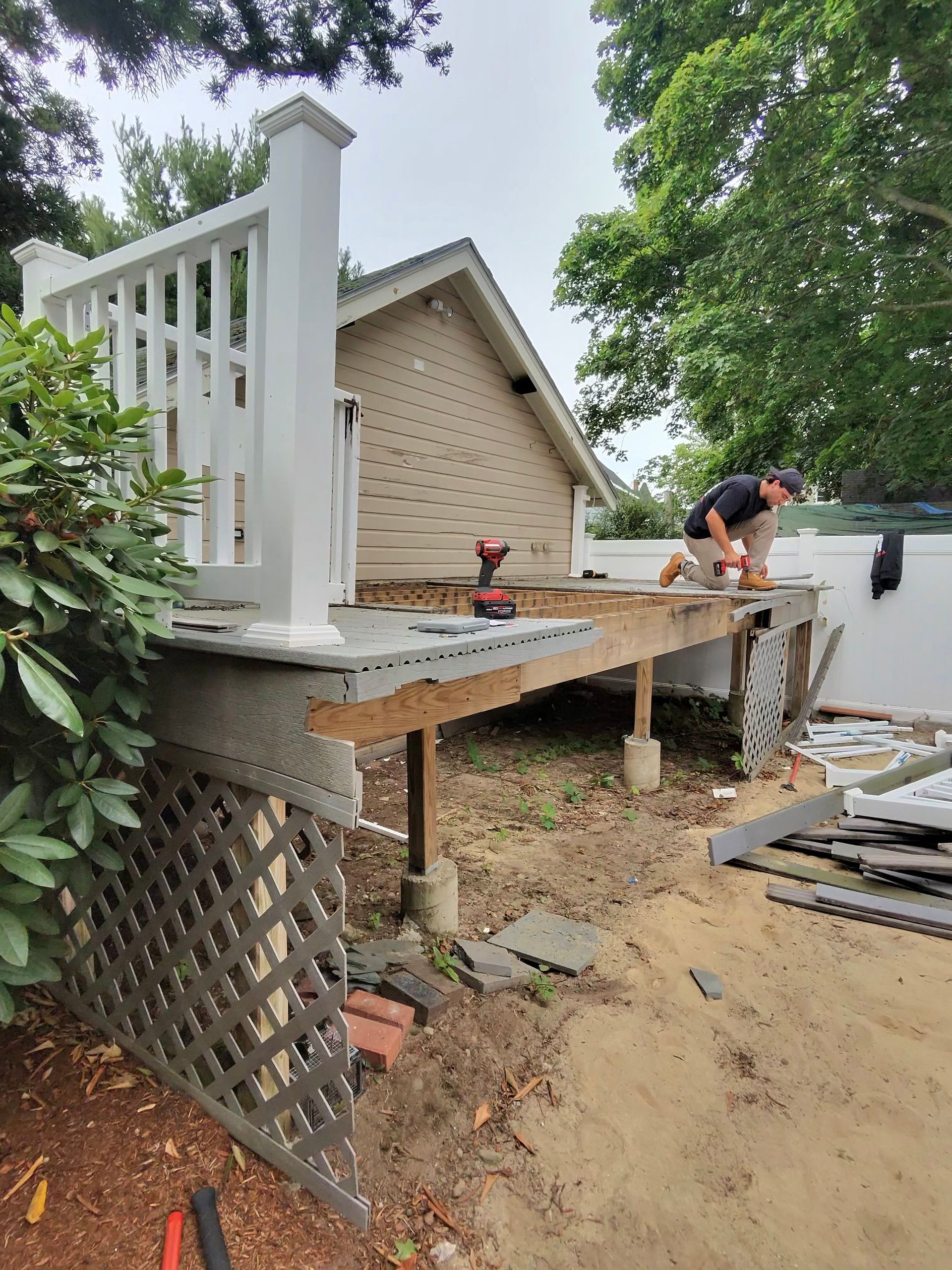 Worker building a raised wooden deck beside a beige house near a sandy lakeside yard
