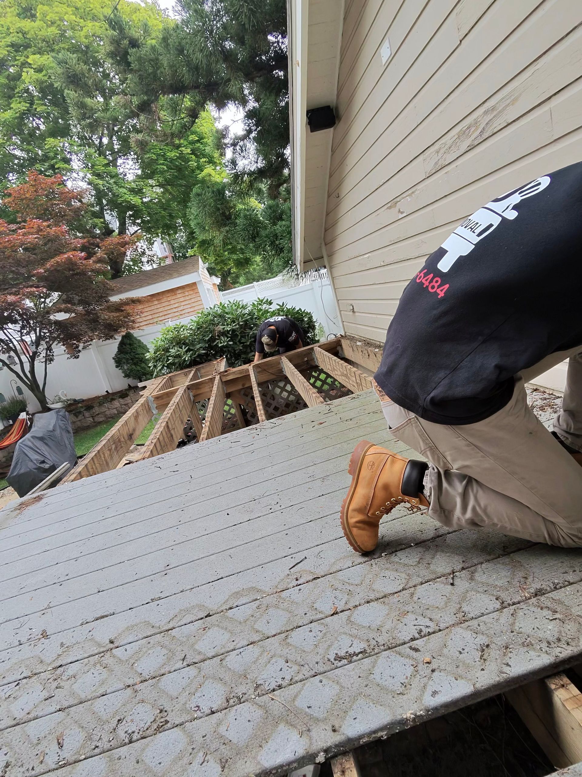 Person kneeling on a slanted deck beside a house, working near a garden with a tool.