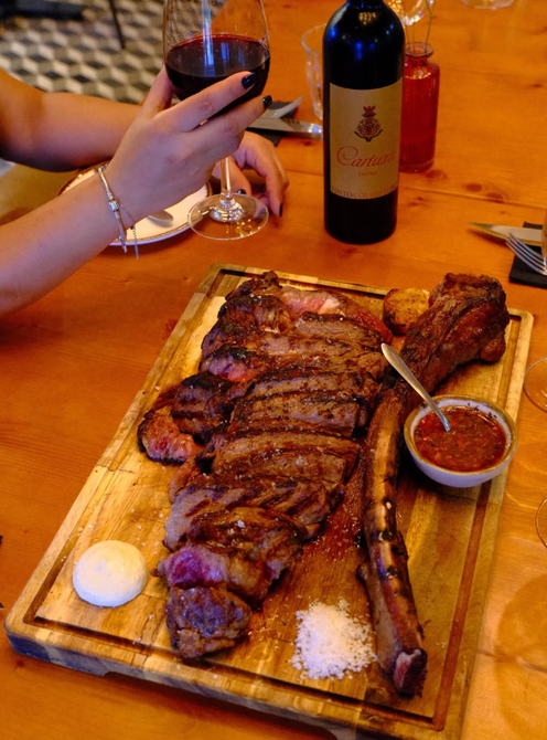Bife grelhado em tábua de madeira com uma taça de vinho tinto, uma garrafa de água e uma tigela de acompanhamentos.