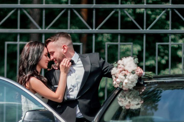 Couple embracing beside a car, with a bouquet visible near the hood and a metal fence in the background